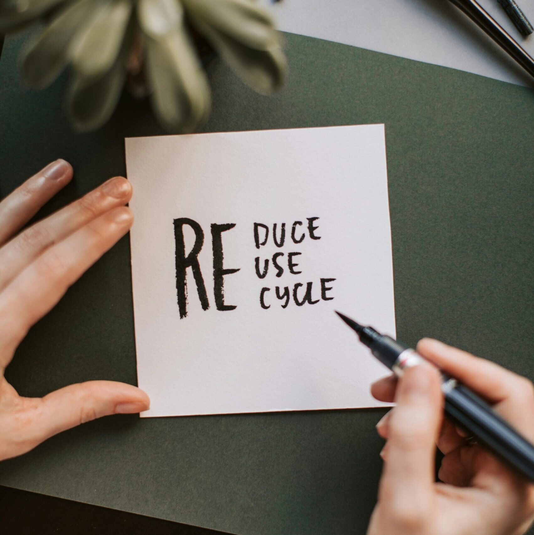 Close-up of hands writing a motivational 'Reduce Reuse Recycle' note on paper with a pen, surrounded by office items.